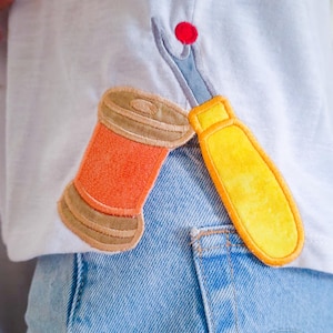 May include: White t-shirt with a colorful embroidered design featuring a spool of orange thread, a yellow seam ripper, and a red pin. The shirt is paired with blue denim jeans.