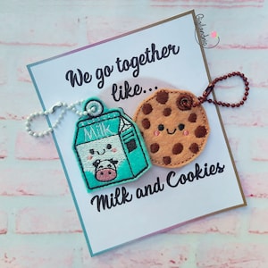 Puede incluir: Una tarjeta blanca con el texto "We go together like... Milk and Cookies" y dos llaveros de fieltro unidos. Un llavero es una caja de leche azul con una vaca sonriente y el otro es una galleta de chocolate marrón con una cara sonriente.