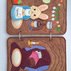 May include: A brown felt book with two metal rings featuring embroidered scenes. The top page shows a rabbit in a blue apron with the text Gigilandnio, a basket of carrots, and a campfire. The bottom page depicts a room with a bed and candle.