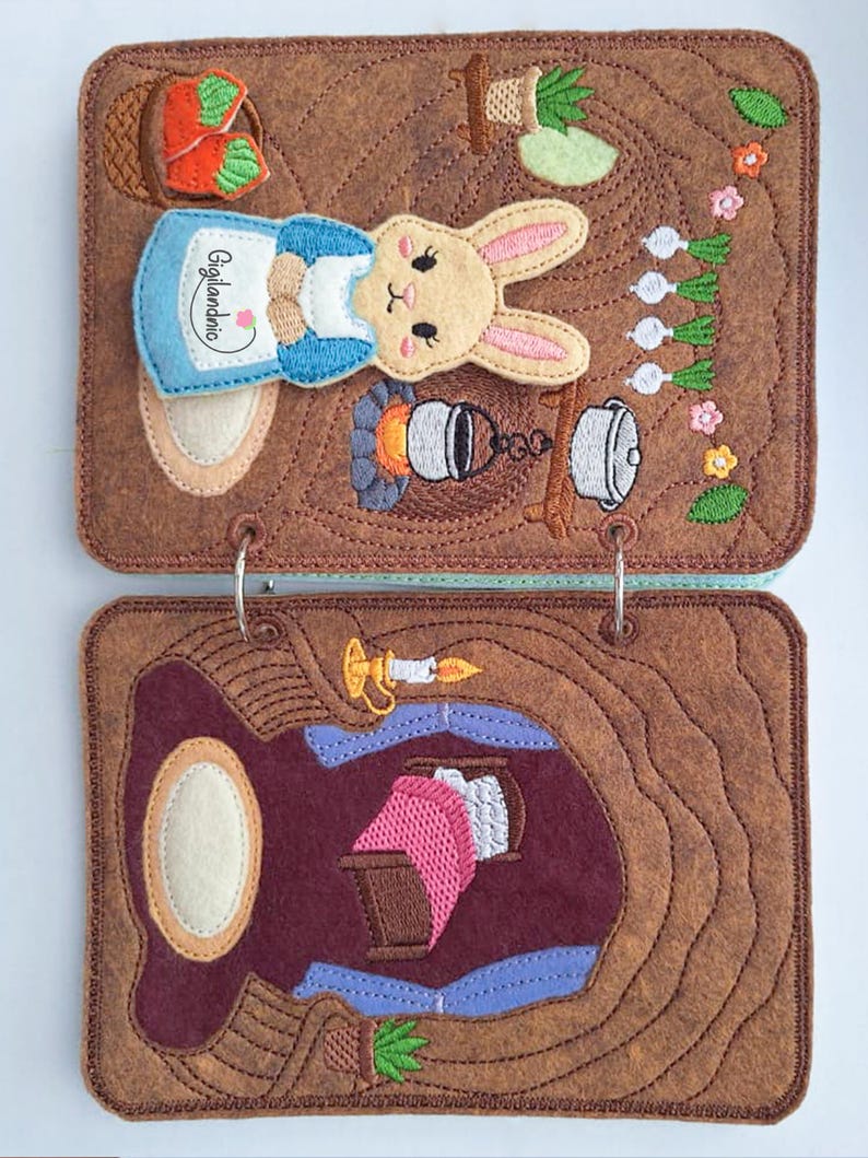 May include: A brown felt book with two metal rings featuring embroidered scenes. The top page shows a rabbit in a blue apron with the text Gigilandnio, a basket of carrots, and a campfire. The bottom page depicts a room with a bed and candle.