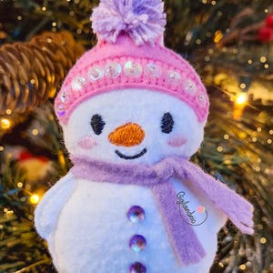 Puede incluir: Adorno navide&ntilde;o de mu&ntilde;eco de nieve con gorro rosa, pomp&oacute;n morado y bufanda. El mu&ntilde;eco de nieve tiene una nariz de zanahoria naranja, ojos negros y tres botones de gemas moradas. El adorno est&aacute; colgado en un &aacute;rbol de Navidad.