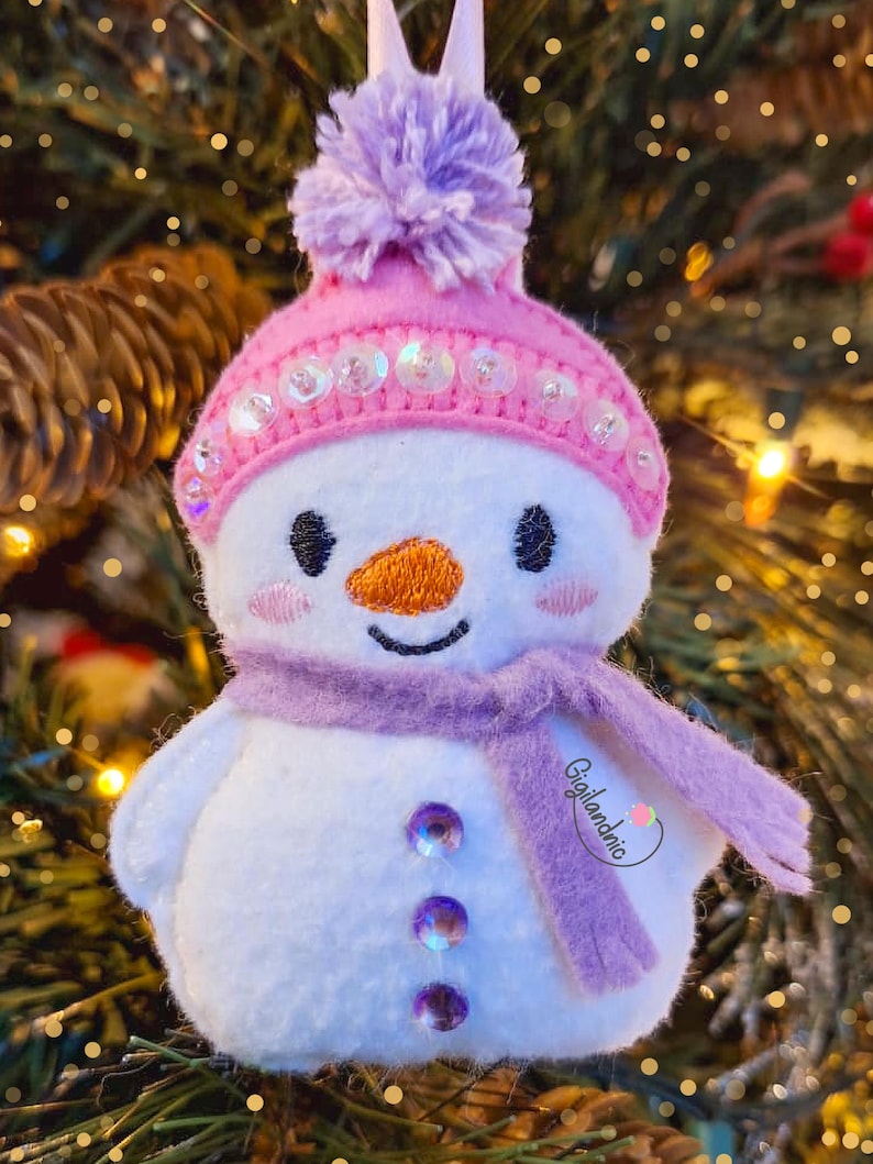 Puede incluir: Adorno navide&ntilde;o de mu&ntilde;eco de nieve con gorro rosa, pomp&oacute;n morado y bufanda. El mu&ntilde;eco de nieve tiene una nariz de zanahoria naranja, ojos negros y tres botones de gemas moradas. El adorno est&aacute; colgado en un &aacute;rbol de Navidad.