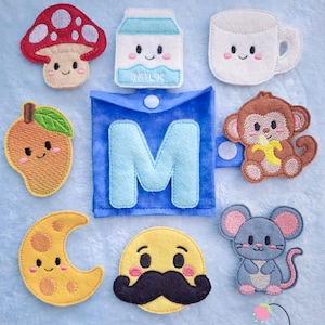May include: A collection of felt embroidered patches featuring a red mushroom, a milk carton, a white mug, a yellow mango, a blue square pouch with the letter M, a brown monkey with a banana, a yellow crescent moon, a yellow face with a moustache, and a grey mouse.