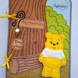 May include: A rectangular felt activity page featuring a brown tree trunk with a door labeled Mr. Sanders, a gold bell, and a sign reading Ring Also. A yellow bear with a white shirt stands on a brown ground next to green foliage.