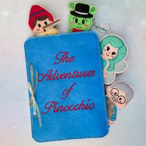 Może przedstawiać: Niebieska książka z filcu z haftowanym tytułem "The Adventures of Pinocchio" wykonanym czerwoną nicią. Książka ma zawiązaną wokół złotą wstążkę i cztery filcowe palczaki wystające z boków. Palczaki to chłopiec w czerwonym kapeluszu, zielony kosmita, syrena i starzec.