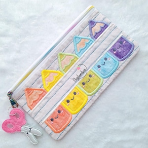 Peut inclure: Une trousse à crayons zippée, ornée de motifs de crayons colorés et de visages souriants. Un charm en feutre rose en forme de ciseaux est suspendu à la fermeture éclair. La trousse a un motif à rayures blanches et grises.