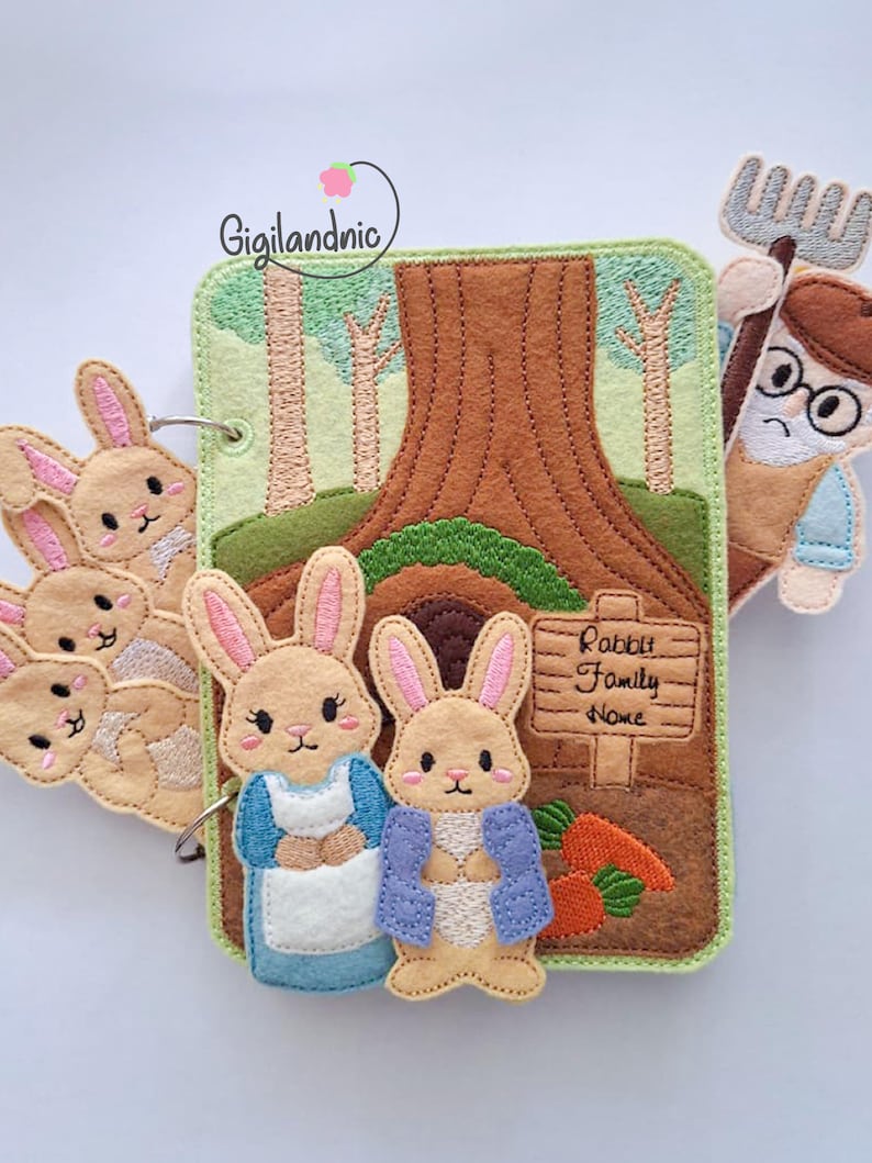 May include: A felt book featuring embroidered rabbit characters, a tree, and a sign reading Rabbit Family Nome. The cover includes two main rabbit figures, a group of three smaller rabbits on a ring, and a separate figure holding a garden tool.