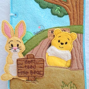 May include: A rectangular felt craft featuring a light blue sky, a brown tree, and green hills. A yellow bear sits near the tree, and a tan rabbit stands beside a brown sign that reads "dont feed the Bear" in dark brown stitched lettering.