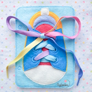 May include: A felt activity board featuring a blue sneaker design with a white toe cap, red tongue, and yellow trim. The shoe is laced with a long, multi-colored ribbon tied in a bow. The rectangular base is light blue with the text Gigilandnic.