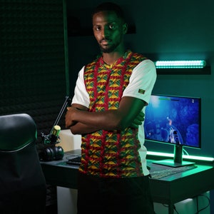 Może przedstawiać: Osoba w białej koszulce z czerwono-żółto-zielonym wzorem. Osoba stoi przed monitorem komputera i biurkiem z mikrofonem.