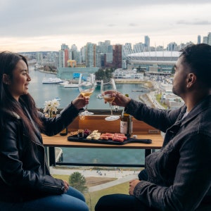 May include: A couple toasting with glasses of white wine on a rooftop patio overlooking a city skyline. They are enjoying a charcuterie board with cheese, meat, and crackers. The table has a sign that says "VIEWS".