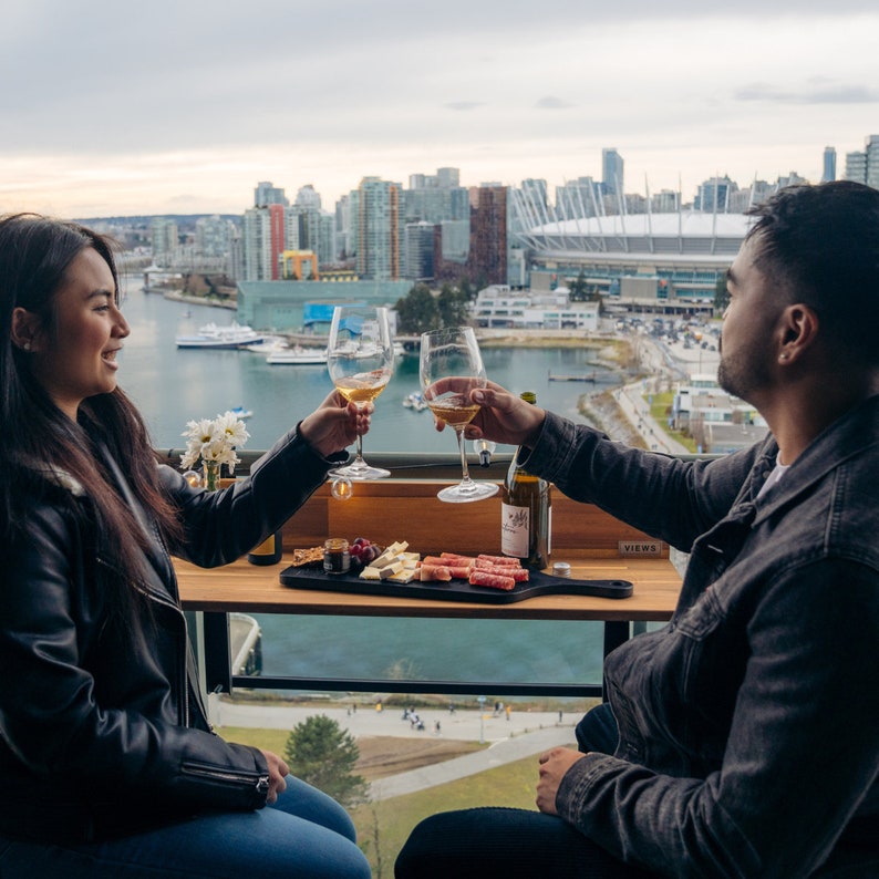 May include: A couple toasting with glasses of white wine on a rooftop patio overlooking a city skyline. They are enjoying a charcuterie board with cheese, meat, and crackers. The table has a sign that says "VIEWS".