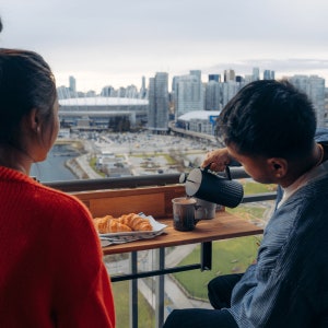May include: A couple enjoys a breakfast of croissants and coffee on a balcony overlooking a city skyline. The man is pouring coffee from a black French press into a white mug. The balcony has a small wooden table with a black metal frame.