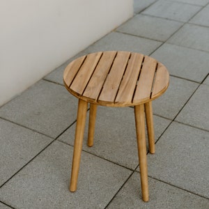 Puede incluir: Una pequeña mesa auxiliar redonda de madera clara, con una tapa de listones y tres patas anguladas. La mesa es de madera clara y está sobre una superficie de baldosas grises. Mide unos 46 cm de diámetro y 46 cm de altura.