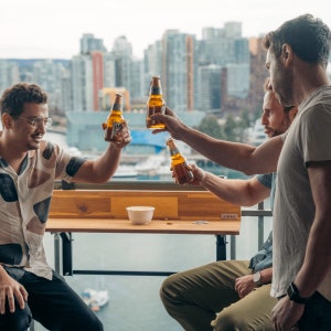 May include: Three friends raise their bottles of beer in a toast on a balcony overlooking a city skyline. The balcony has a wooden railing and a wooden table with a bowl of snacks. The table has a sign that says "VIEWS".