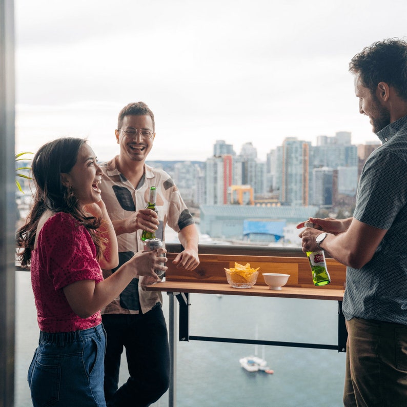 May include: Three people are standing on a balcony with a city view. They are holding bottles of beer and a can of soda. There is a small table with snacks on it.