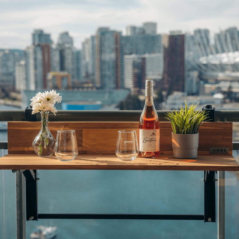 May include: A wooden folding table with a bottle of rose wine, two wine glasses, a vase with white flowers, and a potted plant. The table is labeled "VIEWS" and is mounted to a balcony railing.