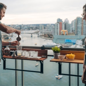 May include: Two people enjoying drinks and snacks on a balcony with a city view. A wooden folding table with the word "VIEWS" printed on it is attached to the railing. The table is holding glasses, a bottle, and a shaker. A second table is holding snacks and a bowl of salsa.