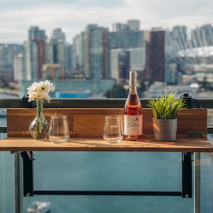 May include: A wooden folding table with a black metal frame, set up on a balcony with a city view. Two wine glasses and a bottle of rose wine are on the table, along with a vase of white flowers. The table has a sign that says "VIEWS".