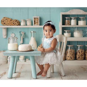 Peut inclure: Une petite fille en robe blanche et bandeau bleu est assise à une petite table bleu clair. Sur la table, un gâteau et un verre de lait. Des étagères en arrière-plan présentent des biscuits et des bocaux d'ingrédients, créant une scène de cuisine charmante.