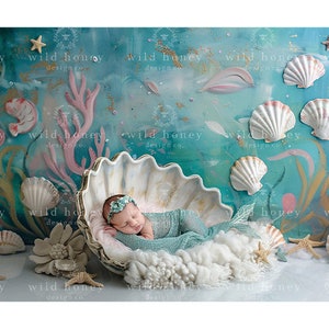 May include: A newborn baby girl is sleeping in a large white seashell prop. The seashell is surrounded by white fluffy fabric and a blue and green mermaid tail. The background is a blue and green ocean-themed backdrop with seashells and coral.