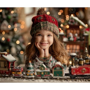 May include: A young girl wearing a red and green knit hat smiles while sitting in front of a toy train set. The train set is decorated with snow and includes a red engine, a green boxcar, and a brown caboose.