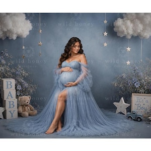 Puede incluir: Un vestido de maternidad azul polvoriento con escote de hombros caídos, mangas largas transparentes y una abertura frontal. El vestido presenta una falda de tul en capas que se extiende por el suelo. Incluye bloques de madera con BABY y un letrero It's a Boy.