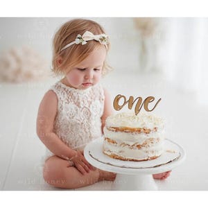 May include: A young child wearing a white lace outfit and a floral headband sits near a small cake on a white cake stand. The cake is topped with a wooden "one" cake topper.