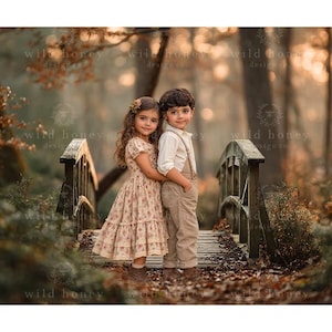 Puede incluir: Un niño pequeño con un vestido de flores de manga corta y falda escalonada está junto a otro niño con una camisa clara de botones y un peto beige sobre un puente de madera en un bosque con hojas de otoño.