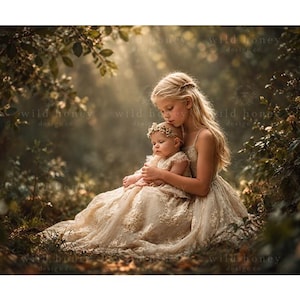 Può includere: Due bambini che indossano abiti in pizzo color crema abbinati con intricati ricami floreali. Il bambino più grande ha lunghi capelli biondi e tiene in braccio il più piccolo, che indossa una delicata fascia floreale, in un bosco soleggiato.