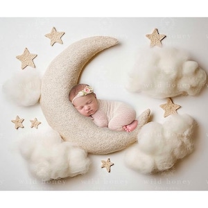 Puede incluir: Un bebé recién nacido durmiendo en una almohada con forma de media luna, rodeado de nubes blancas esponjosas y pequeñas estrellas de madera. El bebé lleva un atuendo de punto blanco y una diadema floral.