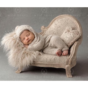 May include: A sleeping infant wearing a beige knit outfit and hat, nestled on a small, ornate, cream-colored chaise lounge. The baby rests on a fluffy, light-colored faux fur blanket. The background is a soft gray.