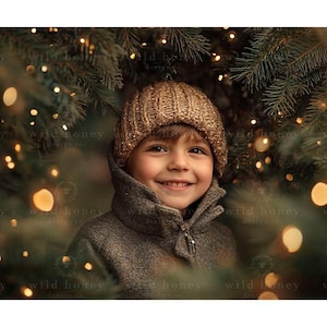 Peut inclure: Un jeune enfant portant un bonnet en laine marron et un manteau marron sourit à la caméra. L'enfant se tient devant un sapin de Noël avec des lumières scintillantes.