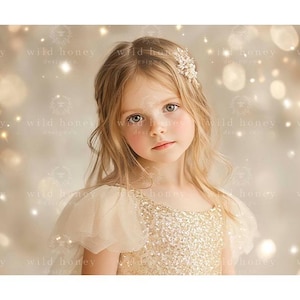 May include: A young girl with blonde hair wearing a gold sequined dress with a white lace overlay and a gold hair clip. The background is a soft, blurred, golden bokeh.