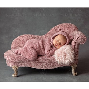 May include: A sleeping baby wearing a pink knit outfit with bear ears, resting on a pink chaise lounge with a floral pattern. A fluffy pink pillow supports the baby's head. The background is a gray backdrop.