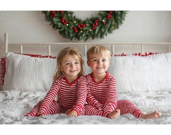 May include: Two children wearing red and white striped pajamas are sitting on a white bed with a white and red patterned blanket. They are smiling at the camera.