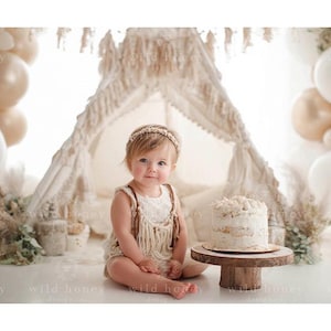 May include: A baby in a cream romper with fringe and a beaded headband sits before a decorative teepee. A small cake on a wooden stand and neutral-toned balloons create a soft, bohemian aesthetic. The scene is set for a special occasion.