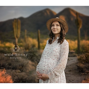 May include: A pregnant woman wearing a white floral dress and a straw hat stands in a desert landscape with mountains and cacti in the background. The text "wild honey design co." is visible in the lower left corner of the image.