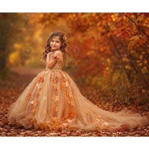 Puede incluir: Un vestido largo hasta el suelo con hombros descubiertos en tonos bronce y dorado, con un corpiño de detalles intrincados y una falda de tul voluminosa adornada con hojas de otoño realistas dispersas en tonos naranja, amarillo y marrón.