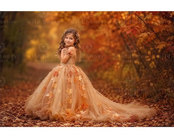 May include: A floor-length, off-the-shoulder gown in shades of tan and gold, featuring a bodice with intricate detailing and a voluminous tulle skirt adorned with scattered, realistic-looking autumn leaves in orange, yellow, and brown tones.