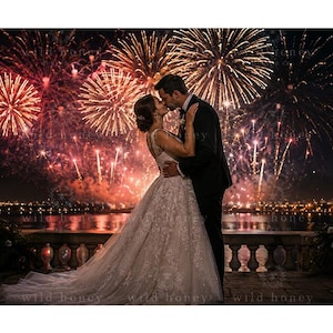 Peut inclure: Un couple en tenue de mariage s'embrassant sur une terrasse en pierre la nuit. La personne à gauche porte une robe en dentelle blanche avec une jupe ample, et la personne à droite porte un costume noir. Des feux d'artifice éclatent dans le ciel.