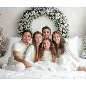 May include: A family of five poses for a Christmas photo in a white room with a snowy Christmas wreath and two snowy Christmas trees. The family is wearing white clothing and smiling.
