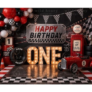 May include: A race car themed birthday backdrop featuring a balloon arch in red, black, and white, a metal sign reading HAPPY BIRTHDAY, large illuminated letters spelling ONE, a red vintage toy car, a fuel pump, and a checkered flag pattern.