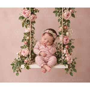 May include: A sleeping infant wearing a light pink long-sleeved romper and a floral headband, sitting on a white wooden swing suspended by ropes. The swing is decorated with lush green foliage and clusters of soft pink roses and baby's breath.