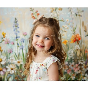 May include: A young girl with long blonde hair smiles at the camera. She is wearing a white dress with a floral pattern. The background is a colorful floral mural.
