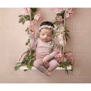 Op de afbeelding: Een baby in een roze gehaakt outfit met een bloemenkrans zit op een schommel versierd met roze bloemen en groen.