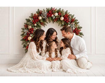 May include: A family of five wearing coordinating white and cream lace dresses and a white button-down shirt with light trousers, positioned in front of a large, semi-circular Christmas wreath decorated with red berries, gold ornaments, and pine.