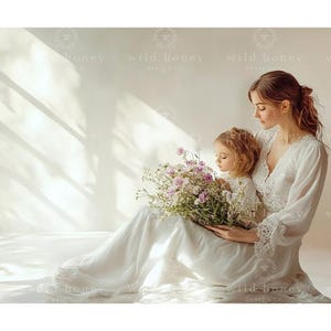 Puede incluir: Una madre y su hija posan juntas en un estudio, ambas vestidas con vestidos blancos. La madre sostiene un ramo de flores blancas y rosas. El fondo es una pared blanca con una ventana que deja entrar la luz natural.