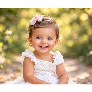 May include: A toddler with brown hair wearing a white lace dress with ruffled sleeves and a light pink hair bow. The child has a smiling expression and is positioned in a natural outdoor setting with soft, blurred green and yellow foliage.