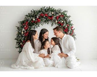 May include: A family of five, dressed in white, smiles in front of a Christmas wreath. The wreath is decorated with green pine, red berries, and gold ornaments. The family is sitting on a white surface.